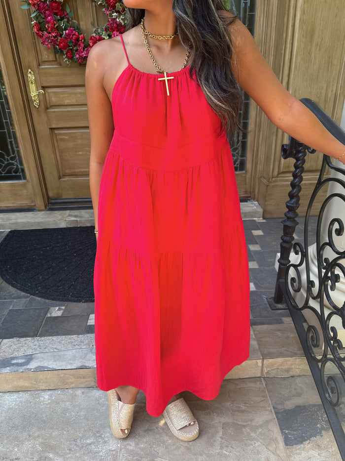 A woman wearing a bright red tiered maxi dress with thin spaghetti straps and a gold cross pendant necklace, standing on a stone patio in front of a wooden door.