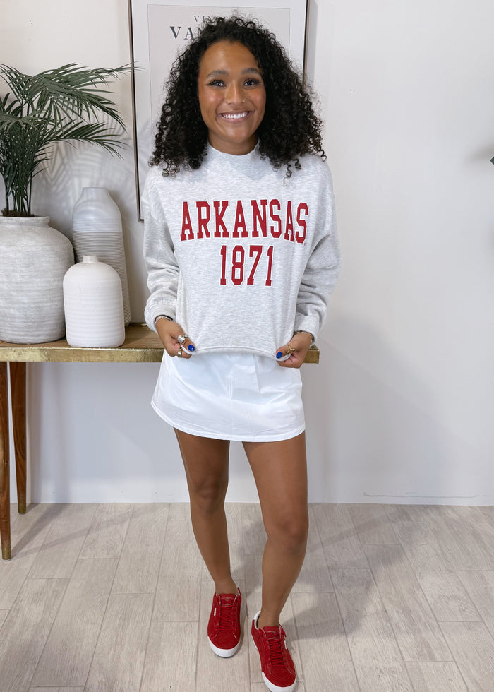 A woman stands indoors wearing a light gray sweatshirt with red 'ARKANSAS 1871' lettering, a white mini skirt, and red sneakers. The room has light wood flooring, white walls, and decorative vases and a plant on a shelf.