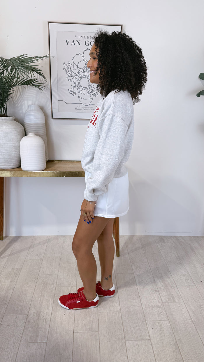 A woman stands in profile wearing a light gray sweatshirt with red lettering, white shorts, and red sneakers in a bright indoor room with wood flooring and wall decor.