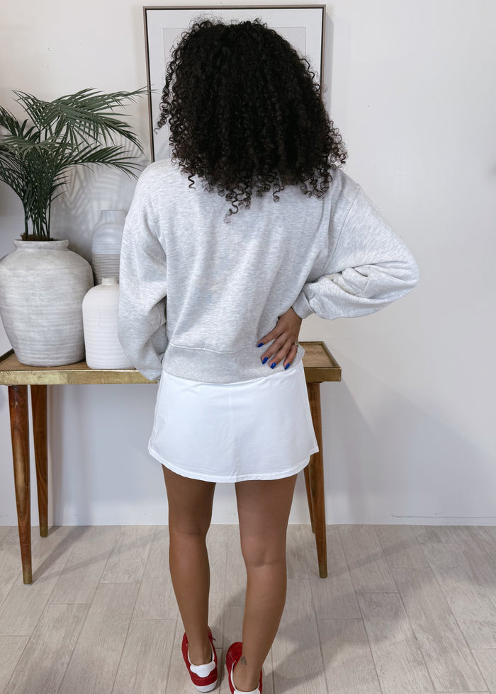 A woman viewed from behind wearing a light gray heathered sweatshirt and a white tennis skirt, standing in a room with light wood floors and a console table.