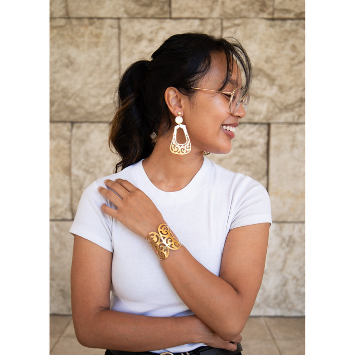 A person wearing the Bactrian Ornament Statement Earrings, which are gold-plated and feature geometric openwork. The earrings are hanging from the person's ears and they are smiling while posing against a stone wall background.