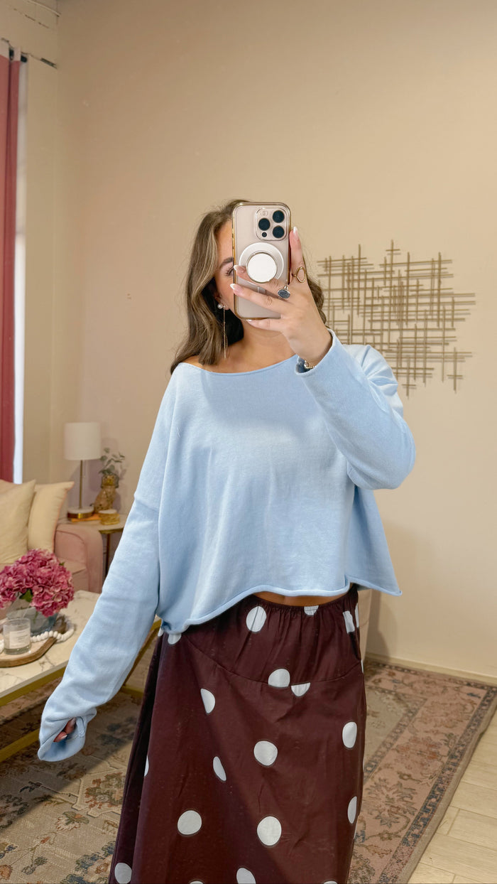 A woman takes a mirror selfie in a room, wearing a light blue long-sleeve cropped top and a dark brown skirt with white polka dots.