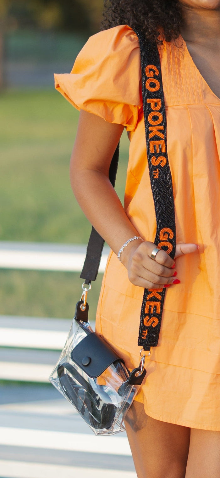 A woman stands outdoors in front of white bleachers and trees, wearing a short orange dress with flutter sleeves and black slide sandals, while holding a small black shoulder bag with orange lettering.