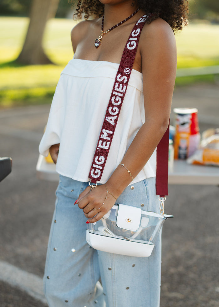 A person outdoors wearing a white off-the-shoulder top, light blue polka-dot jeans, and white sneakers, carrying a clear handbag with a white base and a dark red shoulder strap printed with the text 'GIG EM AGGIES'.
