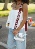 A person outdoors wearing a white off-the-shoulder top, light blue polka-dot jeans, and white sneakers, carrying a clear handbag with a white base and a dark red shoulder strap printed with the text 'GIG EM AGGIES'.