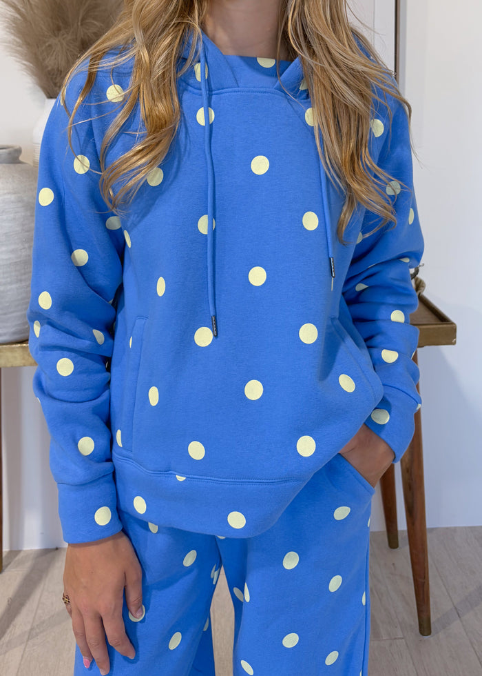 A woman modeling a matching blue hoodie and sweatpants set with a white polka dot pattern, standing in a styled indoor setting with a side table, vase, and framed wall art.