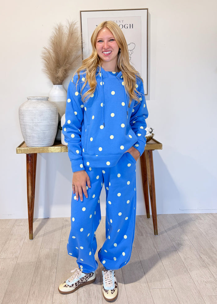 A woman stands indoors modeling a matching blue lounge set with white polka dots, consisting of a sweatshirt and sweatpants, paired with patterned sneakers. She is positioned in front of a light-colored wall with a decorative console table and framed art in the background.
