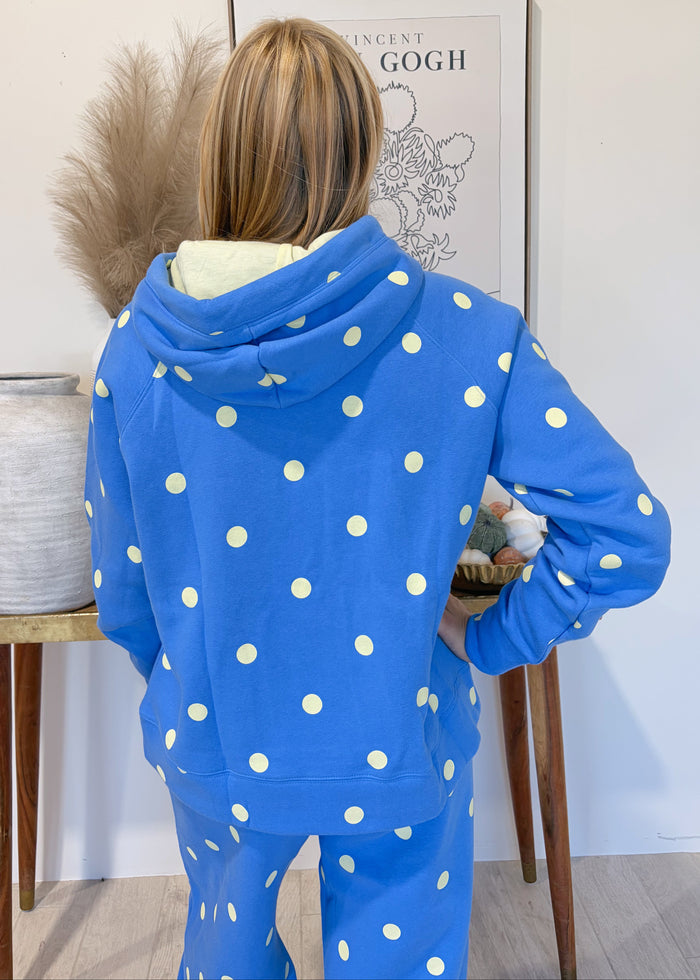 A woman viewed from behind wearing a bright blue matching lounge set with white polka dots, standing indoors in front of a framed print and beside a small table with a vase.