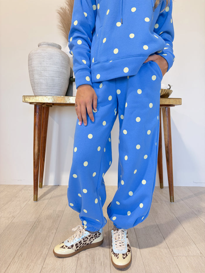 A person wearing a bright blue matching lounge set with white polka dots, including a cropped hoodie and wide-leg sweatpants, paired with leopard-print sneakers, standing in a room with a wooden floor and a side table.