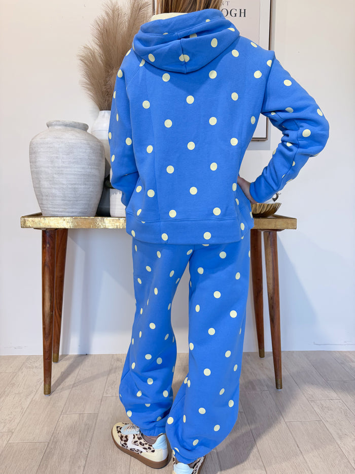 A person wearing a bright blue hooded sweatshirt and matching sweatpants with white polka dots, shown from the back in an indoor room with a wooden table and decorative vase.