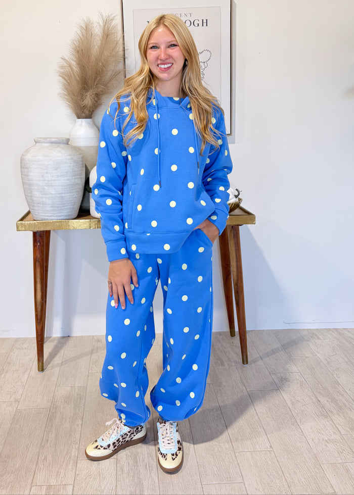 A woman stands indoors modeling a matching blue lounge set with white polka dots, consisting of a sweatshirt and sweatpants, paired with patterned sneakers. She is positioned in front of a light-colored wall with a decorative console table and framed art in the background.