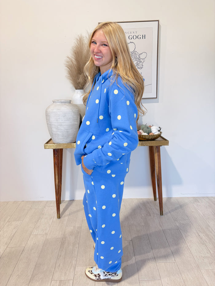 A woman modeling a bright blue one-piece pajama set with white polka dots, standing in a styled room with a wooden console table, a large ceramic vase, and a framed print on the wall.
