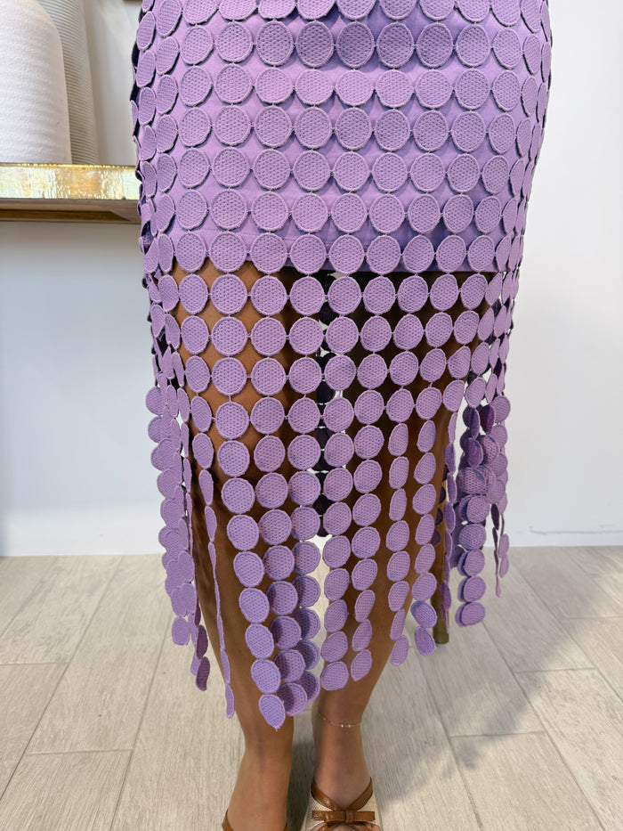 A close-up of a person wearing a lavender purple skirt made of overlapping textured circular cutouts that create a fringed effect, paired with brown strappy heels.