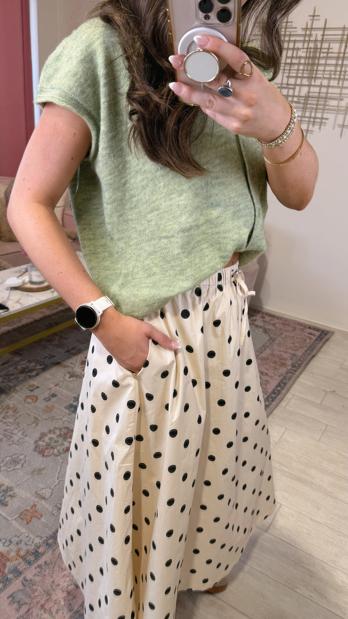 A woman takes a mirror selfie showing an outfit consisting of a light sage green short-sleeve knit top and a cream-colored maxi skirt with black polka dots. She is wearing a white smartwatch and gold jewelry in an indoor home setting.