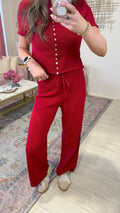 A woman takes a mirror selfie wearing a matching red two-piece set consisting of a short-sleeve button-front top and wide-leg pants. The setting is a home interior with a patterned rug, light wood floor, and a small table with pink flowers.