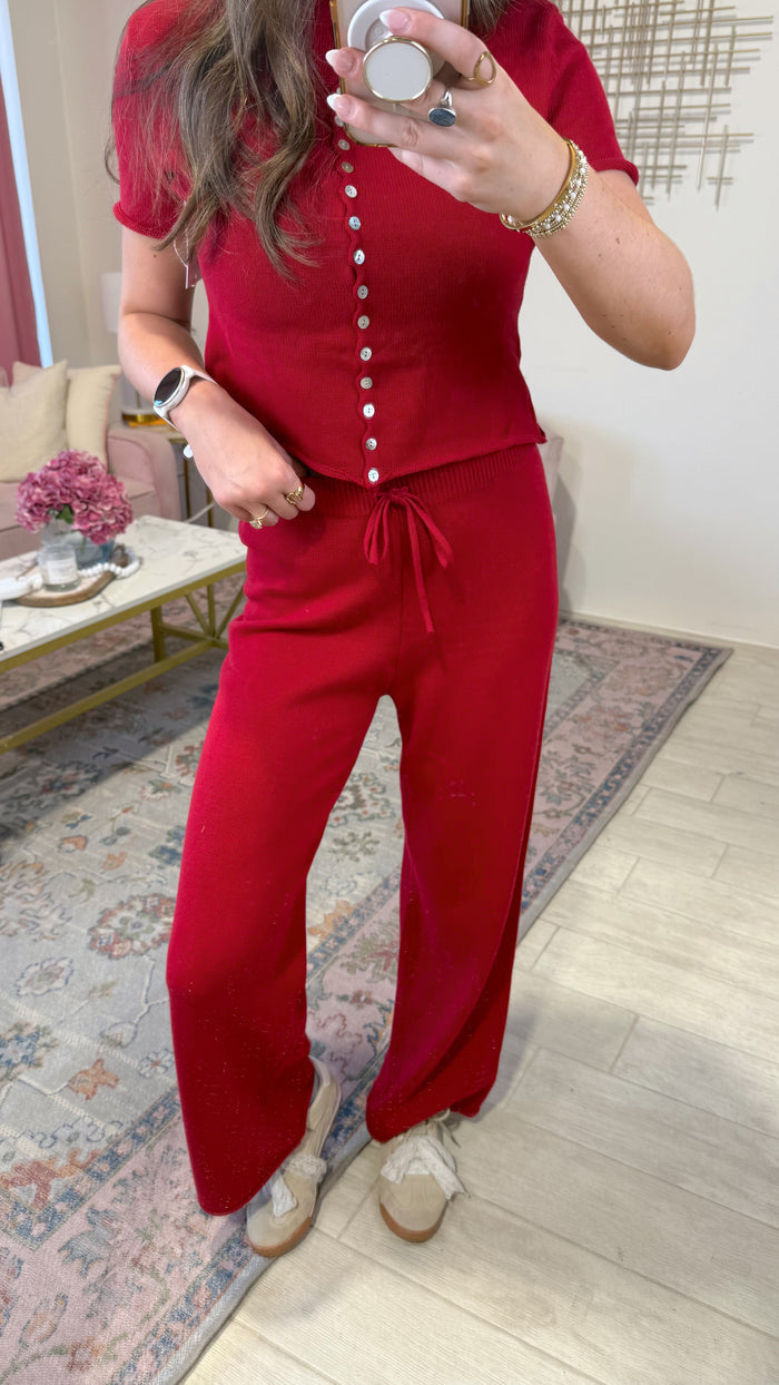 A woman takes a mirror selfie wearing a matching red two-piece set consisting of a short-sleeve button-front top and wide-leg pants. The setting is a home interior with a patterned rug, light wood floor, and a small table with pink flowers.
