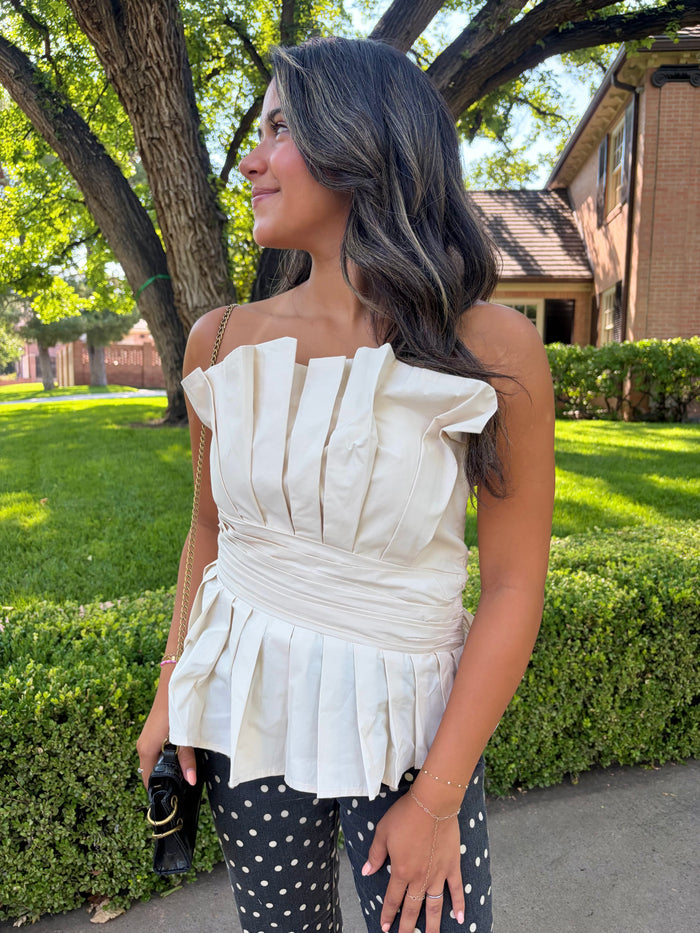 A woman wearing a cream off-the-shoulder ruffled peplum top and black-and-white polka dot pants, standing outdoors in front of a green lawn and trees.