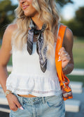 A woman wearing a white sleeveless ruffled crop top, light blue denim shorts, and a black paisley neck scarf, carrying an orange shoulder bag in an outdoor setting.