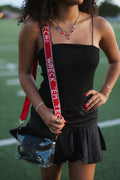 A woman wearing a black spaghetti-strap top and black pleated mini skirt, holding a clear plastic bag with a red and white 'WRECK' strap, standing on a green field.