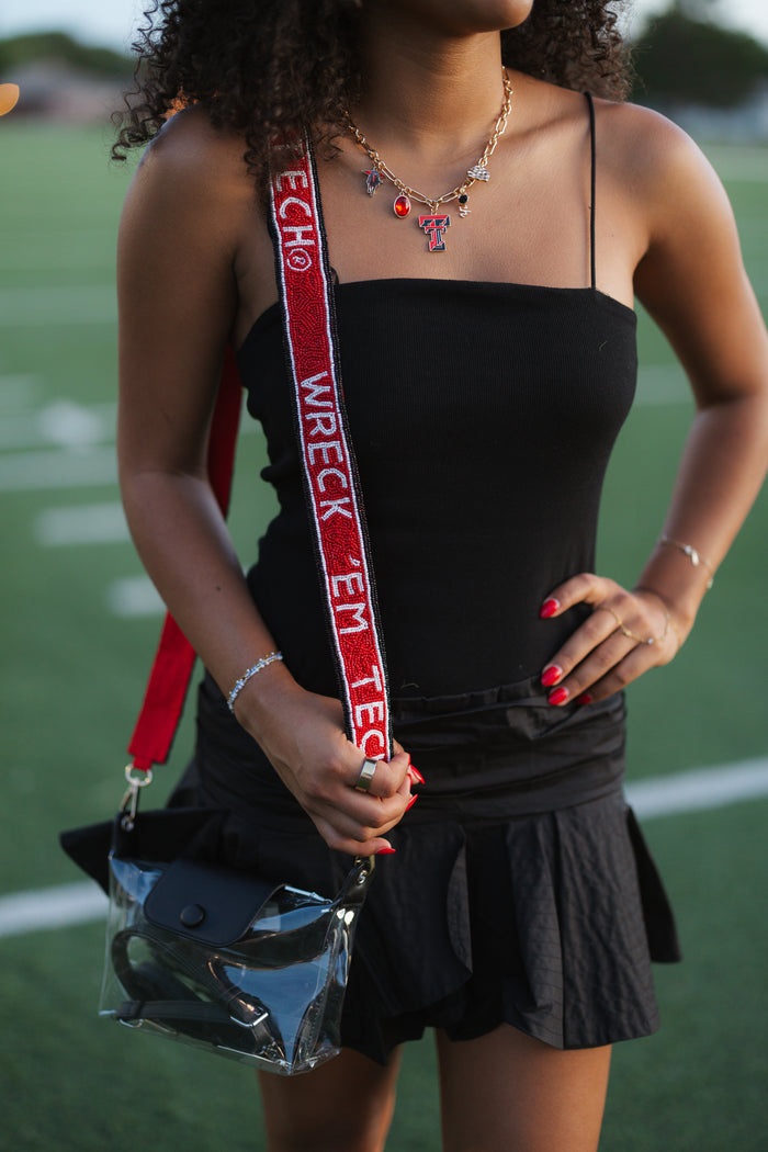 A woman wearing a black spaghetti-strap top and black pleated mini skirt, holding a clear plastic bag with a red and white 'WRECK' strap, standing on a green field.