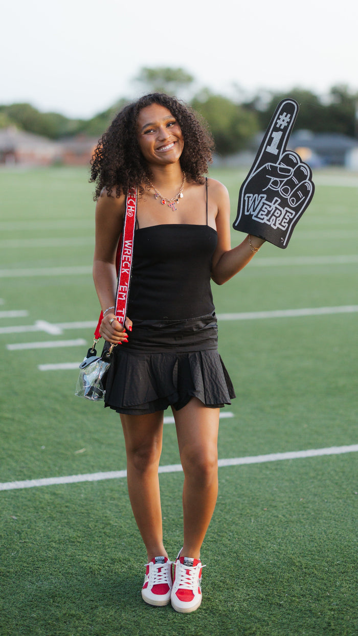 A woman stands on a green football field holding a foam finger and a red-and-white lanyard, wearing a black sleeveless top and pleated mini skirt with white and red sneakers.