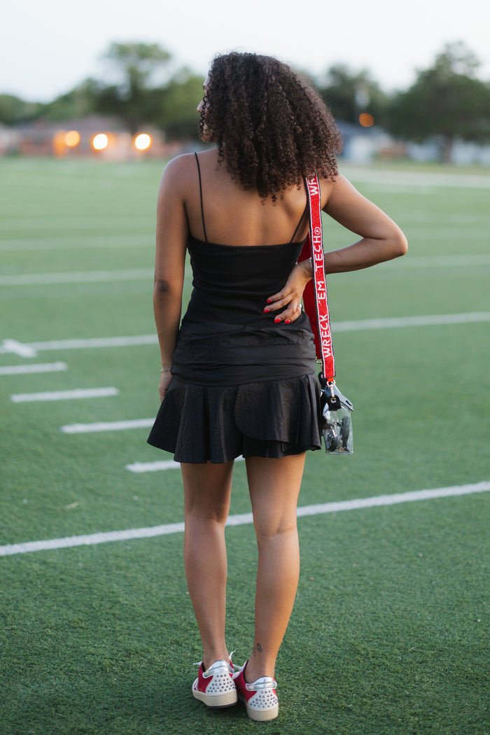 A woman with curly hair stands on a green football field, viewed from behind, wearing a black sleeveless mini dress and holding a red and black shoulder bag.