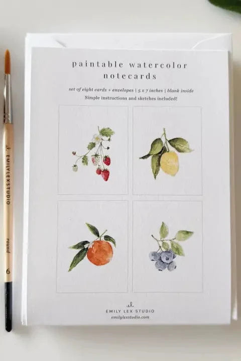 Paintable watercolor notecards with illustrations of fruits and vegetables on a white surface with a paintbrush and fresh produce.
