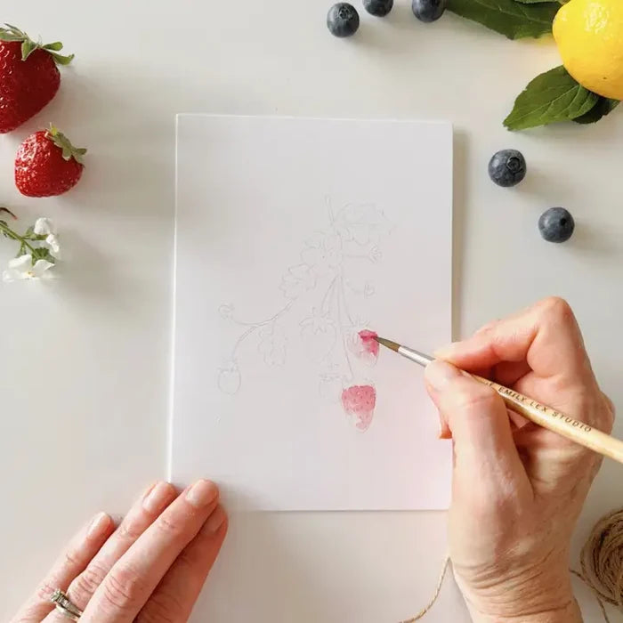 Person painting a strawberry on a white sheet of paper with fruits and flowers around.