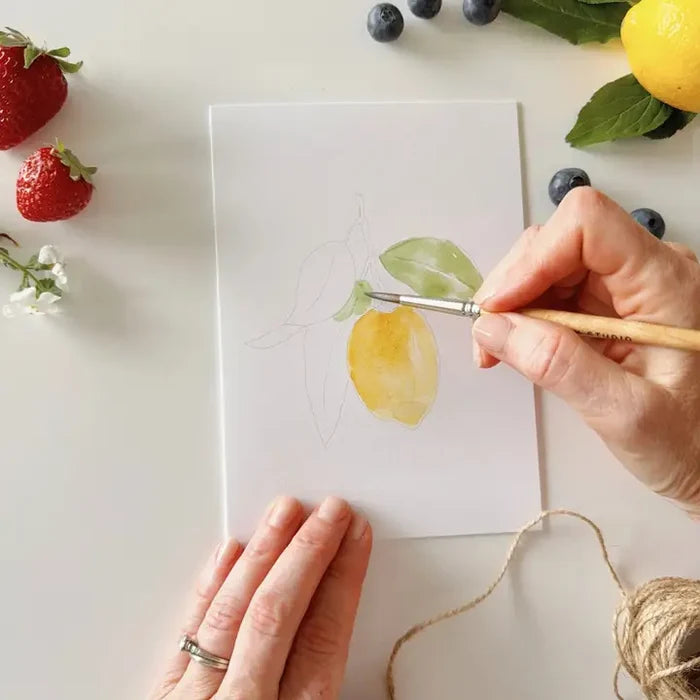 Person painting a lemon on a white sheet with fruits and flowers around