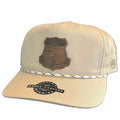 A beige baseball cap with a brown embroidered military-style shield patch on the front, a braided silver cord detail across the brim, and a black circular sticker on the brim.