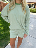 A woman wearing a light sage green and white striped lounge set consisting of a long-sleeve top and matching shorts, standing outdoors on a concrete walkway next to a grassy lawn.
