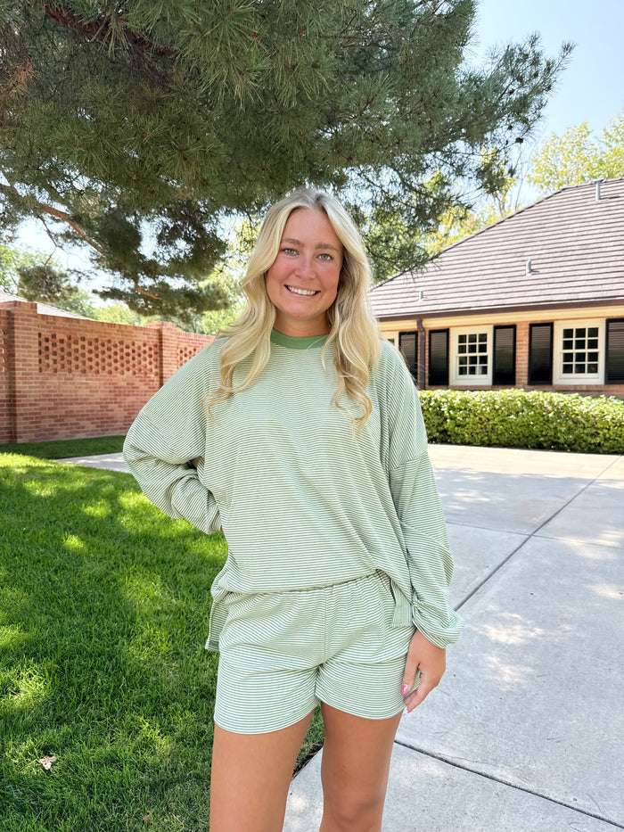 A woman stands outdoors on a driveway in front of a house and tree, wearing a light sage green ribbed long-sleeve top and matching shorts.