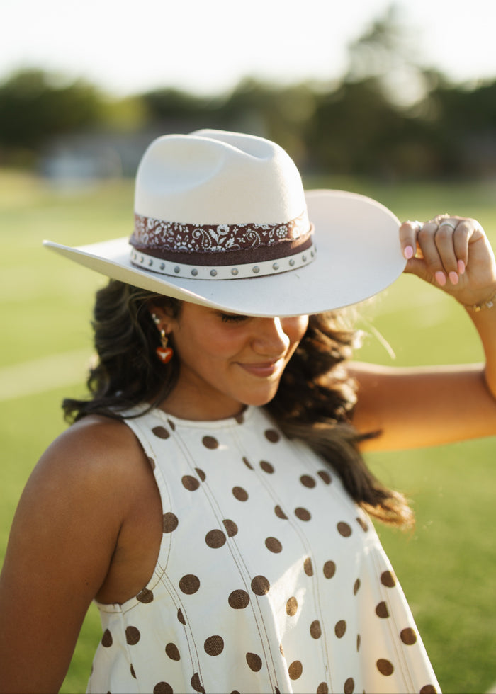 A woman outdoors in a sunny field wearing a white cowboy hat with a brown patterned band and a white sleeveless top with brown polka dots.