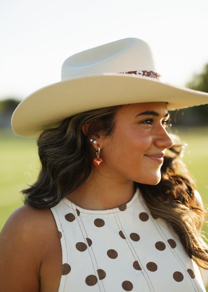 A woman in a white cowboy hat and white sleeveless top with brown polka dots, shown in a warm outdoor portrait with blurred greenery in the background.