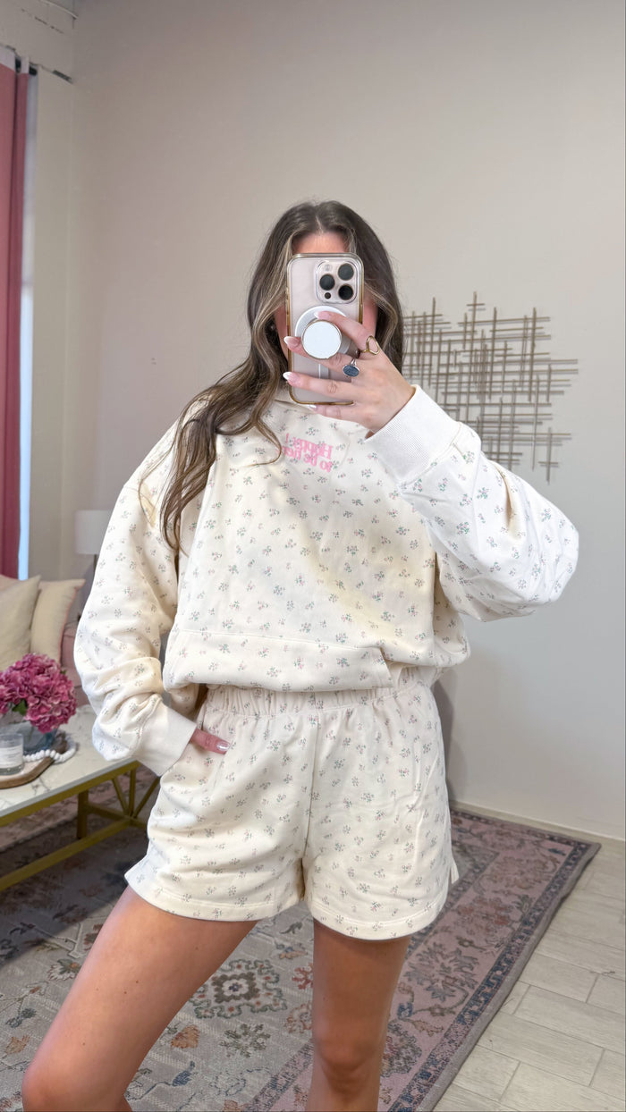 A woman takes a mirror selfie in a room wearing a matching cream-colored lounge set with a small floral pattern. The set includes a long-sleeve sweatshirt and matching shorts.