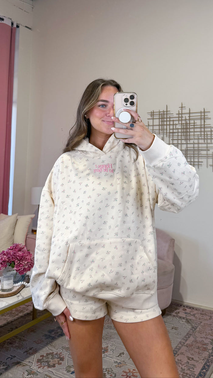 A woman takes a mirror selfie in a room wearing a matching cream-colored loungewear set with tiny pink floral prints. The set includes a pullover hoodie and matching shorts.