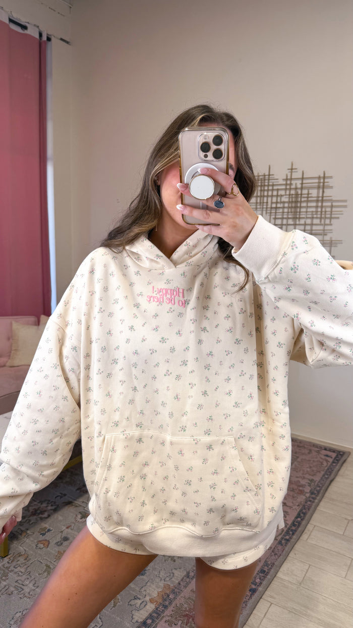A woman takes a mirror selfie wearing an oversized cream-colored hoodie with a small gray floral pattern and pink text on the chest, paired with matching shorts.
