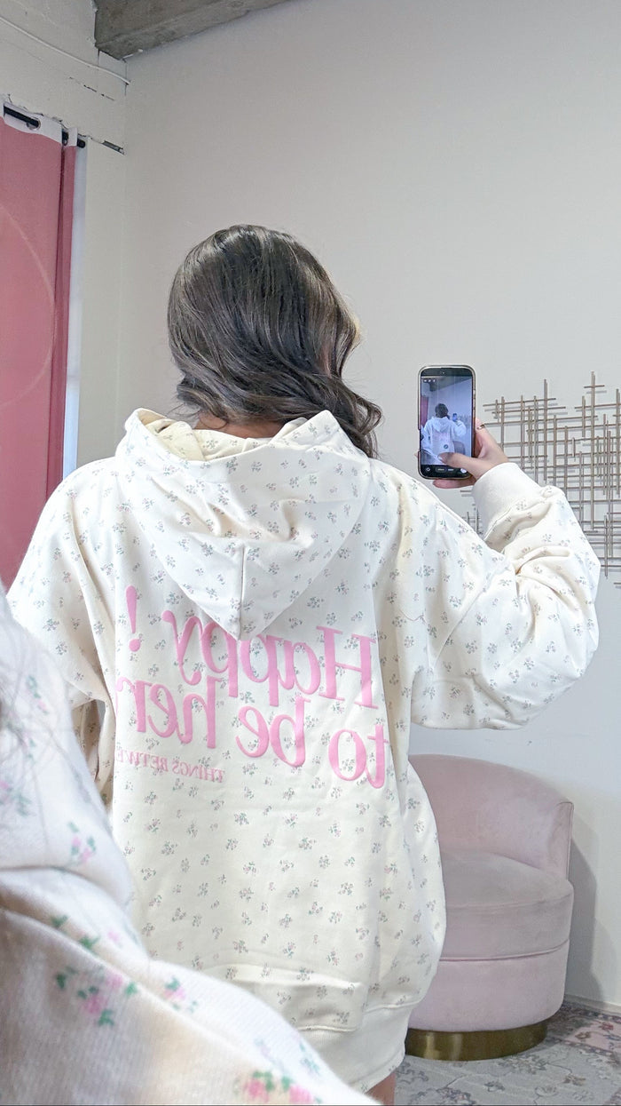 A person viewed from behind wearing a cream-colored hoodie with a tiny floral print and large pink text on the back, holding a phone in a room with a pink chair and white wall.