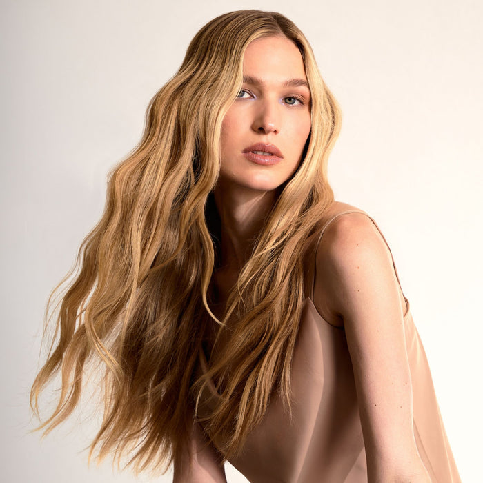 A woman with long, wavy blonde hair wearing a sheer beige satin top, photographed against a clean off-white studio background.