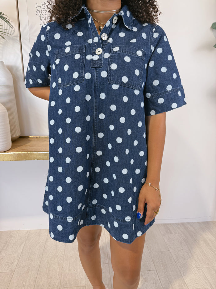 A woman wearing a short-sleeve denim shirt dress with a white polka dot pattern, standing in a brightly lit indoor setting.