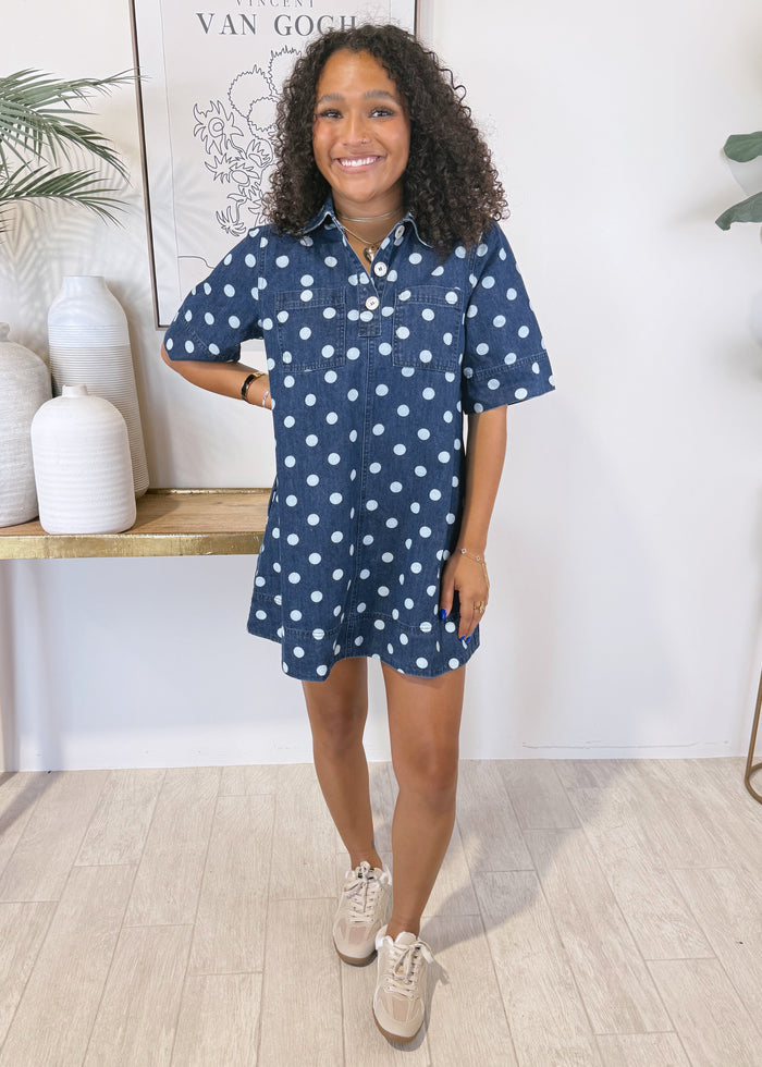 A woman modeling a navy blue short-sleeve shirt dress with white polka dots, styled with beige sneakers in a bright, modern room with light wood floors and decorative vases.