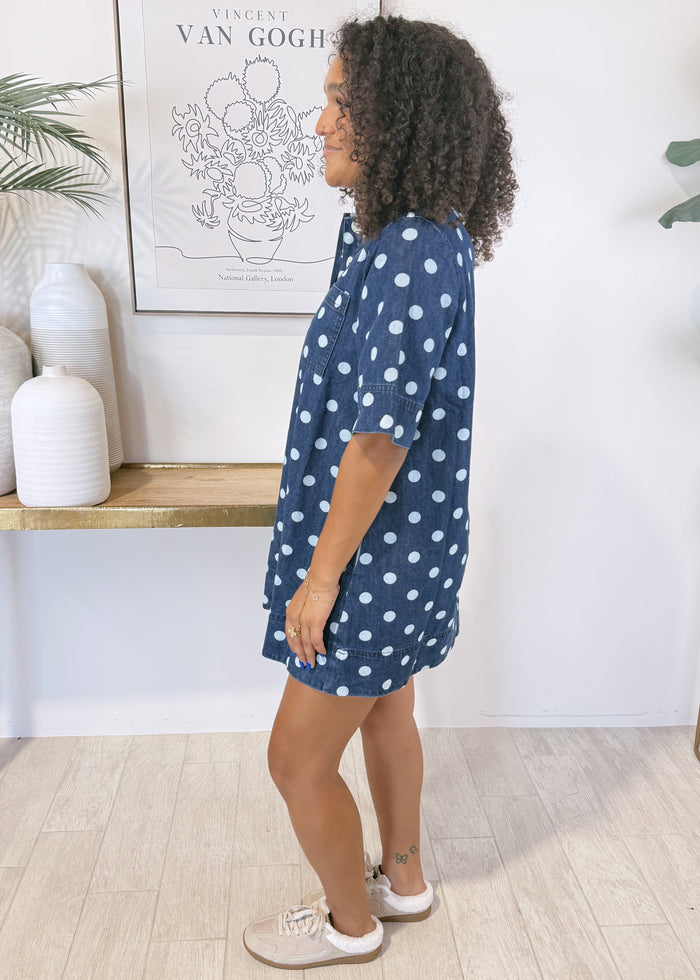 A woman wearing a navy blue short-sleeve button-up shirt dress with white polka dots, standing in profile in a brightly lit room with light wood floors, a console table, and potted plants.