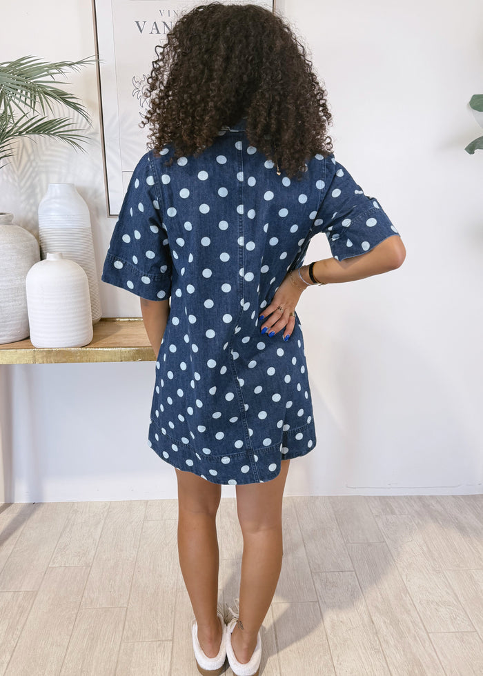 A woman viewed from behind wearing a navy blue short-sleeve shirt dress with white polka dots, standing in a bright room with light wood floors and a console table with decorative vases and plants.