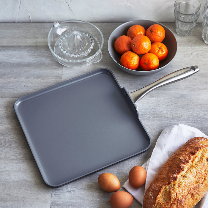 Square ceramic nonstick griddle in gray on a wooden table with eggs, bread, oranges, and a glass juicer.