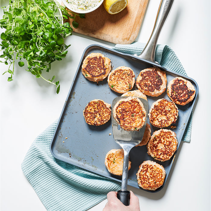 Hand flipping patties on a square ceramic nonstick griddle with greens and lemon; blue textured cloth underneath.