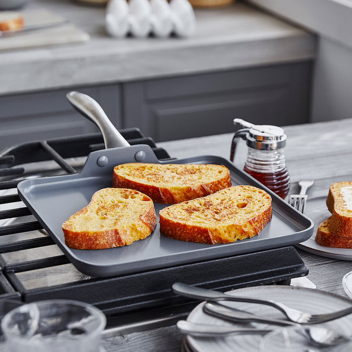 Three slices of French toast on a green ceramic square griddle, with syrup and dishes nearby.
