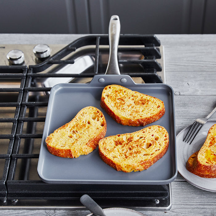 Three slices of bread on a gray ceramic square griddle, with a white plate and silver forks nearby.