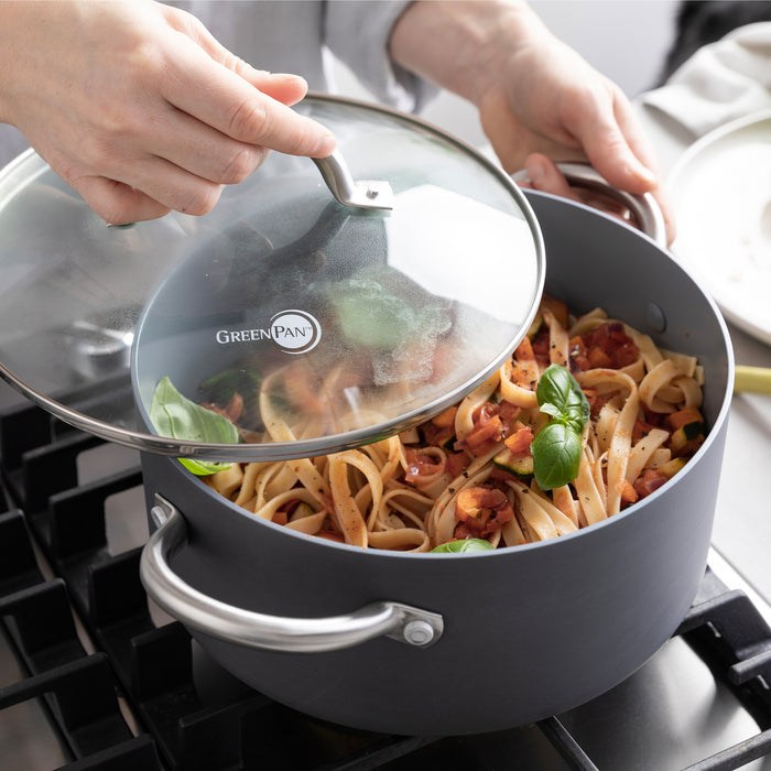 A hand lifts the lid of a gray ceramic nonstick pot with pasta, tomato sauce, and basil on a stovetop.
