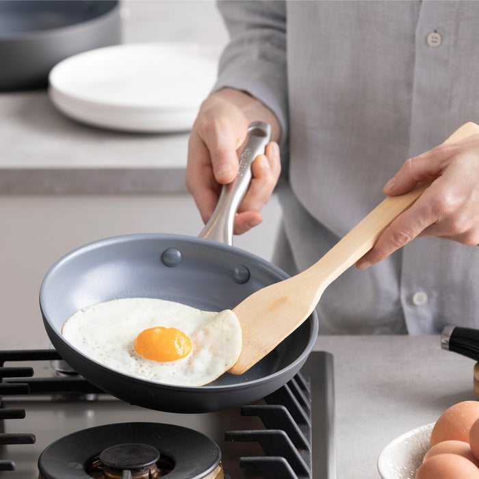 A sunny-side-up egg cooks in a Lima Ceramic Nonstick pan with bamboo utensils on a stove.