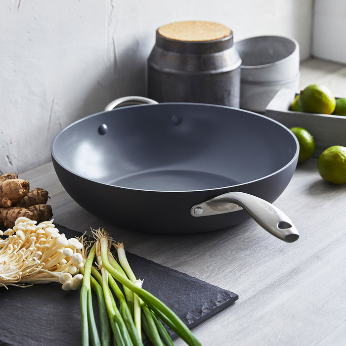 Gray ceramic nonstick wok with green onions, mushrooms, limes, and clear containers on a countertop.